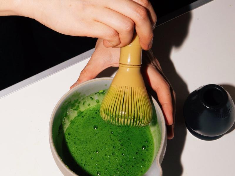 A bamboo chasen whisking matcha to a fine foam in a pale ceramic bowl